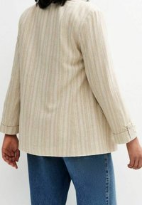 Beige-raitoinen takki, jossa on pitkät hihat ja nappimiehet, valmistettu teksturoidusta kankaasta. Yhdistetty sinisiin farkkuihin. Hieman huomaamaton kuviointi.