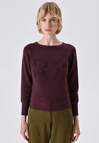 Maglione knit bordeaux con design testurizzato a forma di "R", collo a giro, polsini e fondo a coste. Abbinato a pantaloni su misura verde oliva.
