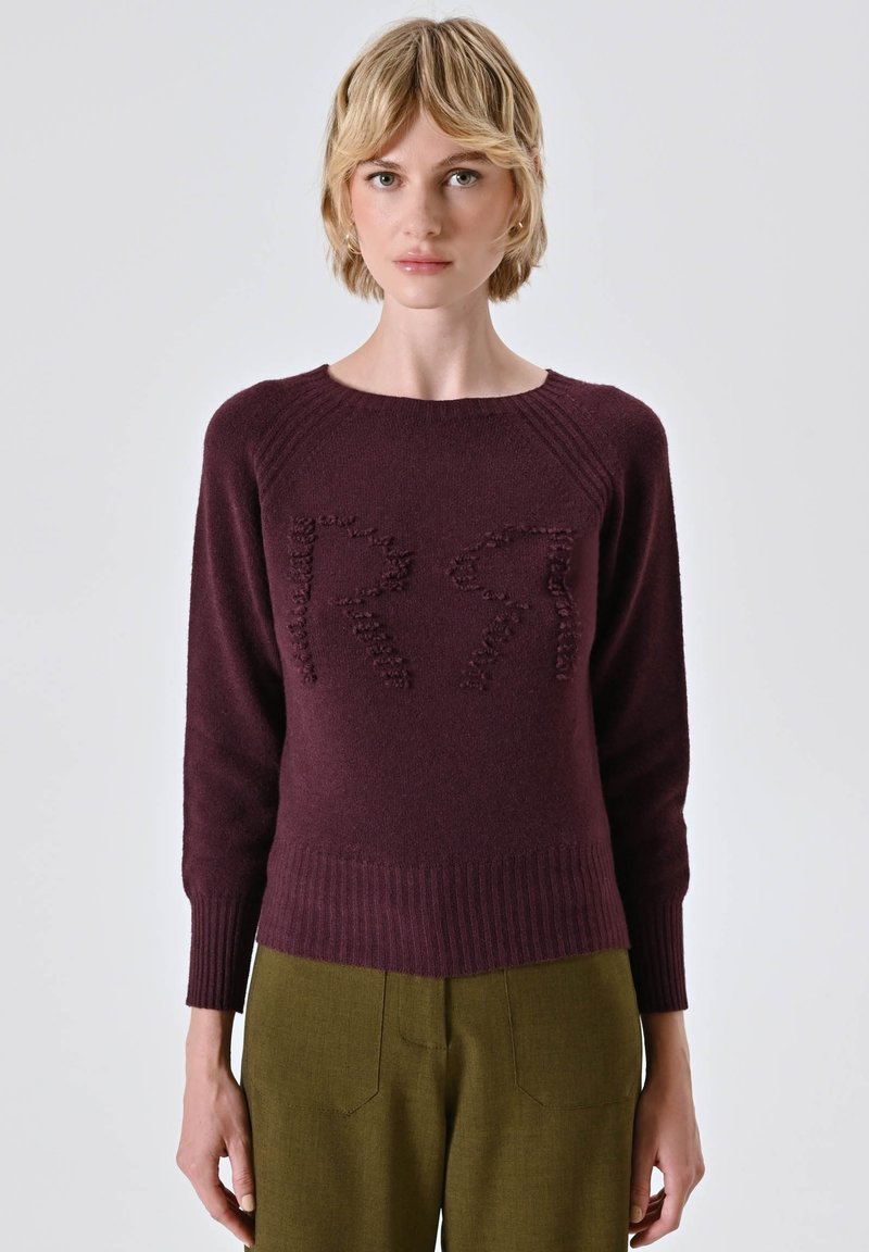 Maglione knit bordeaux con design testurizzato a forma di "R", collo a giro, polsini e fondo a coste. Abbinato a pantaloni su misura verde oliva.