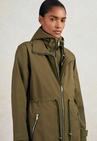 Veste imperméable vert olive avec un col montant, fermeture éclair à l'avant, taille ajustable par cordon de serrage et poches zippées. Présente une texture lisse et un design décontracté.