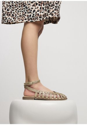 Leg wearing gold glitter strappy sandal with ankle buckle, standing on white rounded pedestal, leopard print skirt hem visible above knee.