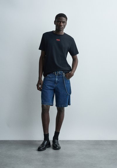 Άνδρας στέκεται μπροστά σε ανοιχτόχρωμο τοίχο φορώντας μαύρο t-shirt GCDS, μπλε τζιν σορτς με ζώνη και αλυσίδα, μαύρες κάλτσες και μαύρα μοκασίνια.