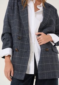 Blazer de cuadros de lana azul marino de doble botonadura, hecho de tela texturizada, con grandes botones marrones, combinado con una camisa blanca y jeans oscuros.