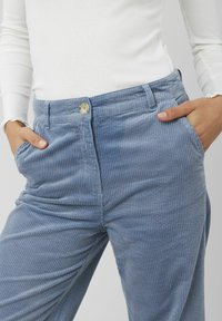 Pantalons en velours côtelé bleu avec des côtes verticales, dotés d'une fermeture à bouton à l'avant et de poches latérales, associés à un haut à manches longues blanc.