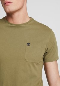 Olijfgroene katoenen T-shirt met een ronde hals, korte mouwen en een patch pocket met een subtiel logo op de bovenste linkerzijde.