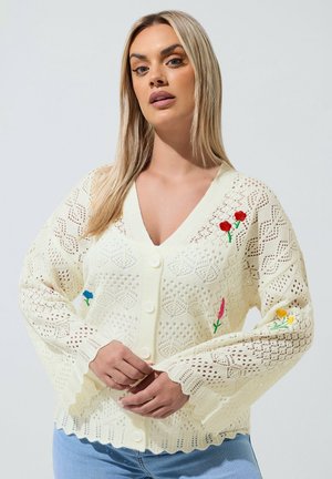 Kvinde iført en cremefarvet strikket cardigan med blomsterbroderi og bølgekant, kombineret med lyseblå jeans.