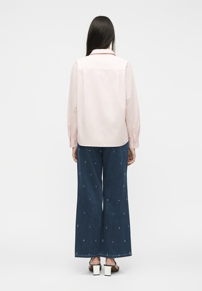 Γυναίκα που στέκεται και έχει την πλάτη στραμμένη, φοράει ένα ανοιχτό ροζ μακρυμάνικο πουκάμισο, σκούρα μπλε κεντημένα τζιν με φαρδιά πόδια και ψηλοτάκουνα με μαύρα λουράκια.