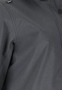 Dunkelgrauer Softshell-Jackenstoff mit sichtbaren Nähten, schwarzem Druckknopf und einem kleinen Ravusco-Markenetikett in der Nähe des Kragens.