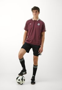 Maillot de football rayé bordeaux et blanc avec un col rond, shorts noirs, chaussures de football noires, chaussettes noires montantes avec des rayures blanches, et un ballon de football.