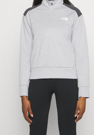 Person iført en lysegrå kvart-zip sweatshirt med sorte skulderpaneler og sorte leggings, stående med armene afslappet ned langs siden.