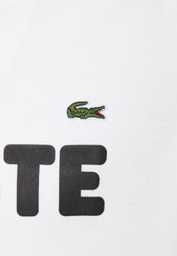Petit logo de crocodile brodé en vert au-dessus de grandes lettres noires "TE" sur un tissu blanc texturé.