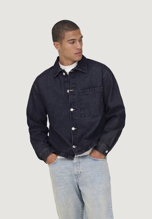 Uomo con giacca in denim scuro con bottoni e jeans azzurri chiari, mani nelle tasche, guarda di lato.