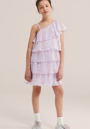 Young girl wearing a sleeveless, lilac tiered dress with silver dots, white socks, and white sneakers, standing against a plain background.