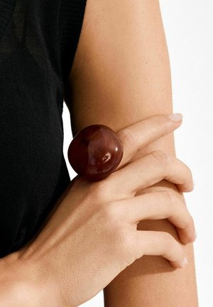 Sjajni, sferični prsten bordo boje od smole nošen na prstu, u kontrastu s golom rukom i crnim tkaninom. Glatka tekstura, velika veličina.