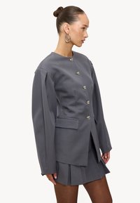 Blazer gris sur mesure avec manches longues, six boutons métalliques, une poche simple, associé à une jupe plissée assortie. Texture lisse.