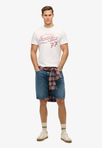 Hvid t-shirt med rød grafisk print, blå denim shorts og en ternet skjorte bundet om taljen. Beigesneakers og lyse sokker.