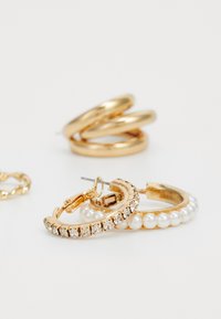 Brincos de argola dourados, apresentando um par com strass transparentes e pérolas, ao lado de tr�ês argolas douradas lisas empilhadas ao fundo.