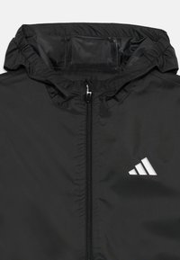 Veste coupe-vent noire avec un zip à l'avant, capuche élastique et logo blanc sur la poitrine. Tissu lisse et léger.