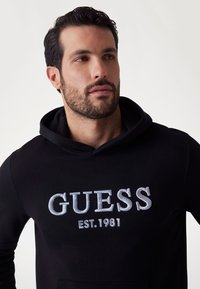 Sudadera negra de tejido suave con un gran logotipo blanco bordado "GUESS EST.1981" en el frente y una capucha amplia.