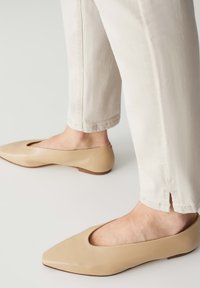 Ballerines en cuir beige avec un bout pointu et des côtés découpés. Semelle plate et texture lisse. Portées avec un pantalon court de couleur claire.