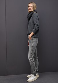 Suéter de punto gris oscuro combinado con jeans grises que presentan una costura lateral decorativa, complementado por zapatillas blancas. Los accesorios incluyen una bufanda negra.