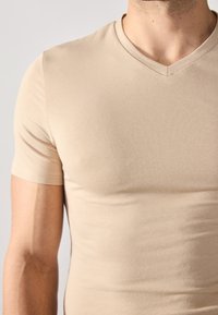 Tricou scurt cu mâneci, de culoare bej, fabricat dintr-un material moale și elastic. Prezintă un decolteu în V și o formă ajustată, fără modele sau logo-uri.