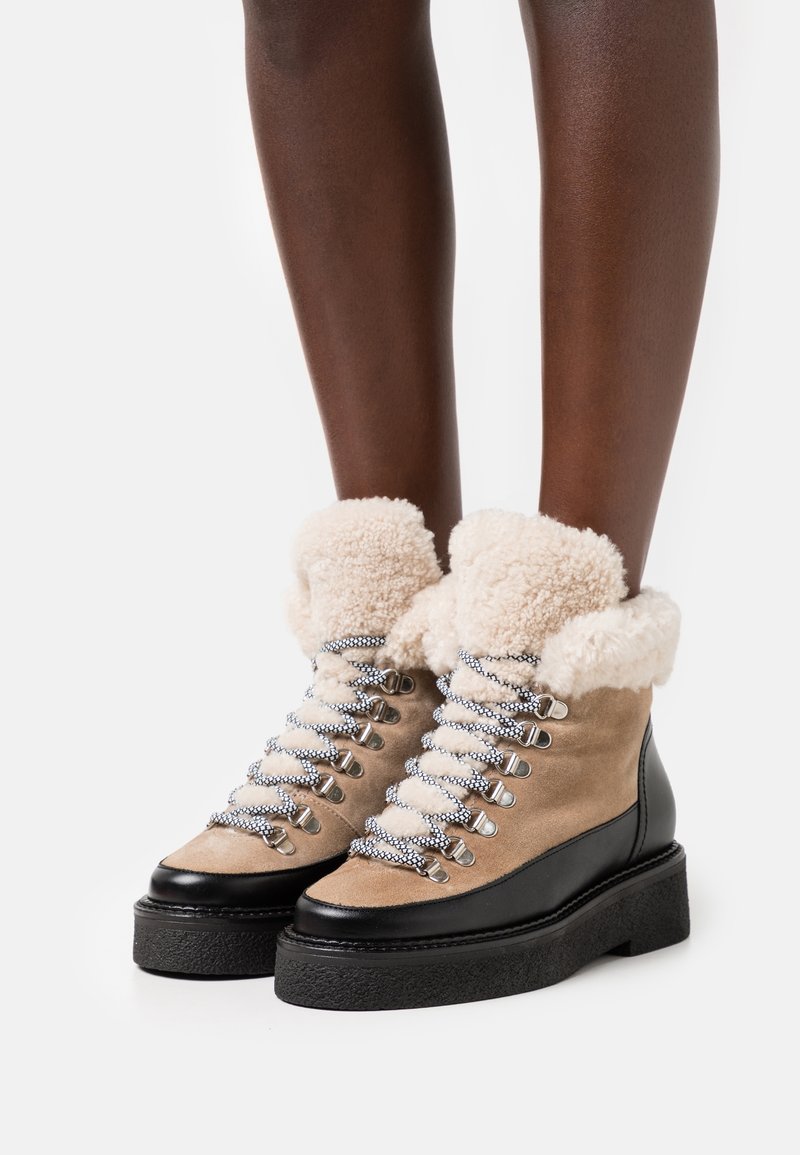 Bottes de cheville en daim beige avec une semelle noire en plateforme, un col en fourrure crème, des éléments en métal argenté et des lacets à motif.