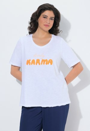 Kobieta z kręconymi włosami do ramion, ubrana w białą koszulkę z napisem "lovely KARMA" w pomarańczowym kolorze i ciemnoniebieskie spodnie, stojąca z rękami za plecami.