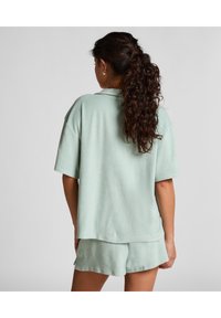 Vrouw met krullend haar in een paardenstaart, gekleed in een lichtgroene badstoffen top met korte mouwen en bijpassende shorts, staand met haar rug naar de camera tegen een witte achtergrond.
