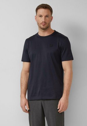 Mężczyzna o krótkich włosach, ubrany w gładki, ciemnoniebieski t-shirt i szare spodnie, stojący na tle jasnoszarego, jednolitego tła.