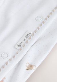 Vita babykläder med beige kant, tryckknappar, ett litet djurtryck och en etikett med texten "Born in 2026."