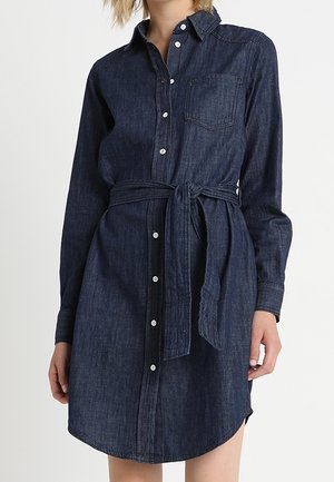 Mujer que lleva un vestido camiseo de mezclilla azul oscuro con mangas largas, botones en la parte delantera, bolsillo en el pecho y un cinturón de tela atado a la cintura.