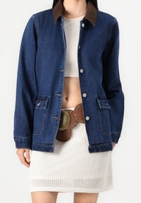 Jeansjacka med en krage av brunt kordfloret, framknäppning med tryckknappar och två sidofickor. Bärs över en vit stickad kjol och ett bälte med cirkulära detaljer.