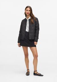 Melna, kviltota jaka ar apaļu kaklu, ar pogu aizdari, apvienota ar baltu kreklu un melnu svārku. Modelis valkā melna līmeņa apavus ar siksnām.