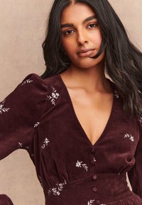 Abito di velluto a coste color bordeaux con maniche a sbuffo, scollo a V e ricamo floreale. Presenta una chiusura con bottoni e un tessuto strutturato per un dettaglio in più.