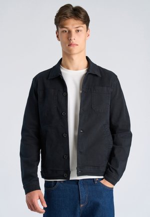 Nuori mies seisoo, päällään musta napillinen takki valkoisen t-paidan ja sinisten farkkujen päällä, yksinkertaisen vaalean taustan edessä.