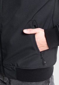 Giacca nera con zip, caratterizzata da una texture liscia, dotata di una tasca laterale e polsini a costine; design minimalista senza fantasie o decorazioni.