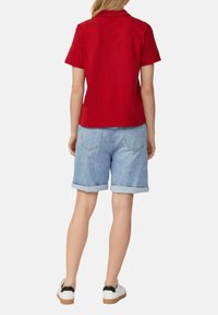 T-shirt polo rouge à manches courtes, associé à un short en jean bleu clair avec les bords retroussés. Le modèle porte des baskets blanches avec des accents noirs.