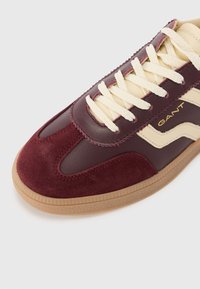 Chaussure bordeaux avec textures en cuir lisse et daim, lacets crème, semelle en caoutchouc et logo latéral. Présente un design athlétique rétro et un bout arrondi.