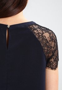 Robe marine avec des manches courtes en dentelle, dotée d'une fermeture à dos en trou de serrure et de détails en bouton métallique. Tissu lisse avec une texture de dentelle complexe.