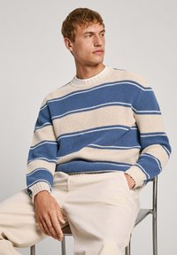 Kootud sviiter, millel on horisontaalsed sinised ja beežid triibud, soonikkoes kaelus ja varrukad, koos heledate pükstega.