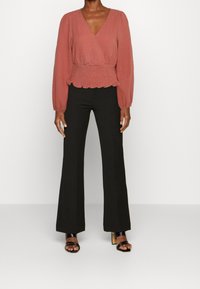 Blusa avvolgente color ruggine con maniche lunghe, vita arricciata e orlo scallop, abbinata a pantaloni neri a palazzo e sandali neri con cinturini.
