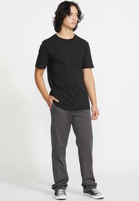 Black cotton t-shirt with short sleeves, round neckline, relaxed fit, paired with gray pants and black sneakers, standing against a white background.