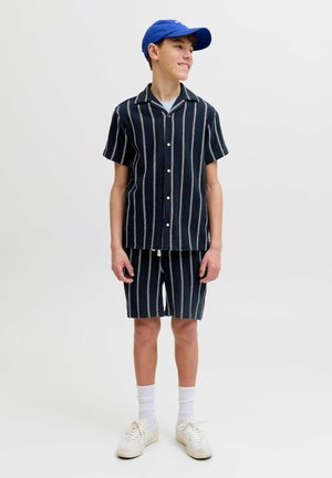 Adolescent purtând un tricou cu mâneci scurte cu dungi verticale alb-negru și pantaloni scurți asortați, adidași albi și o șapcă albastră.