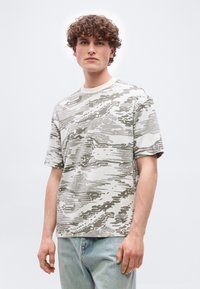 Νέος άνδρας με σγουρά μαλλιά, που φοράει μια άνετη κοντομάνικη γκρι και λευκή μπλούζα με σχέδια και ανοιχτόχρωμα τζιν, στέκεται μπροστά σε μια απλή φόντο.