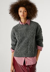 Pelēks adīts džemperis ar teksturētu apdari, kas uzlikts virs sarkana un balta svītrainā krekla, papildināts ar bordo ādas svārkiem.