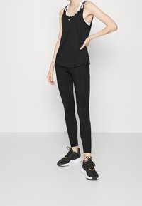 Schwarzes sportliches Tanktop mit Rundhalsausschnitt, kombiniert mit engen schwarzen Leggings mit Nahtdetails und schwarzen Sneakers mit gelben Akzenten.