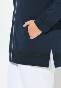 Veste zippée bleu marine en tissu doux, dotée de poches latérales et d'un ourlet légèrement ouvert, complétant un pantalon blanc.