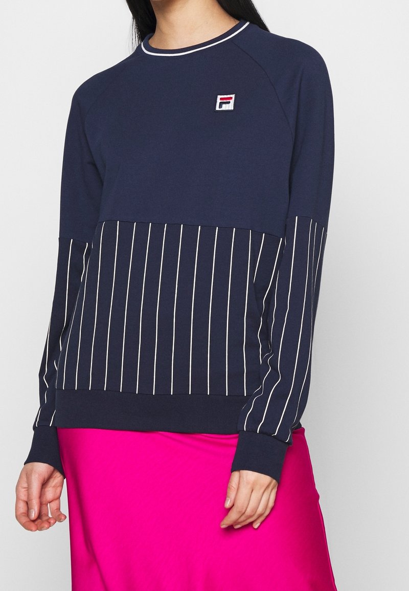 Femme portant un sweat-shirt bleu marine avec des rayures blanches verticales sur les manches et le corps, un petit logo sur la poitrine, associé à une jupe rose vif.