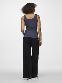 Femme aux cheveux bouclés portant un haut sans manches bleu marine, un pantalon noir à jambes larges et des baskets noires, debout de dos sur un fond blanc.